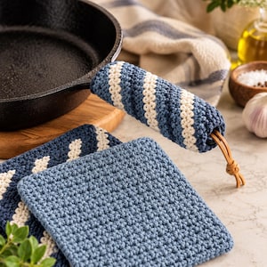 May include: A cast iron skillet handle cover and a square potholder, both crocheted with a blue and cream striped pattern. The handle cover features a tan leather cord tie at the end, while the potholder has a solid blue textured surface.