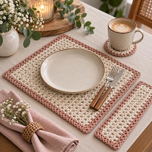 May include: A rectangular crochet placemat and a matching rectangular coaster, both featuring a cream-colored center with a dusty rose border. The set is crafted with a textured stitch pattern and is displayed with a ceramic plate, a fork, and a knife.