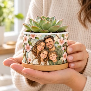 May include: A cylindrical ceramic planter featuring a printed illustration of a family of four surrounded by pink cherry blossoms. The planter sits on a circular bamboo saucer and contains a green succulent plant with thick leaves.