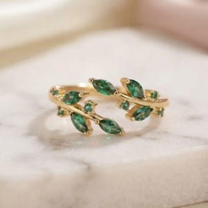 Peut inclure: Une bague ouverte en métal doré présentant un motif de branche avec des pierres vertes taille marquise disposées en forme de feuilles. Les petites gemmes vertes sont serties le long de la structure délicate rappelant une vigne.