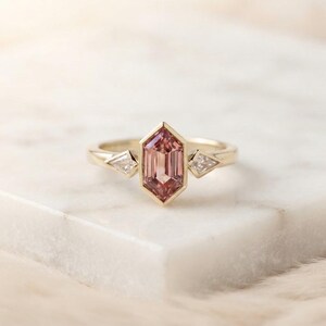 Pode incluir: Um anel de ouro com uma pedra preciosa rosa hexagonal central cravada num bisel, ladeada por duas pedras claras mais pequenas em forma de papagaio de cada lado. A banda é feita de ouro amarelo polido, criando um visual elegante e refinado.