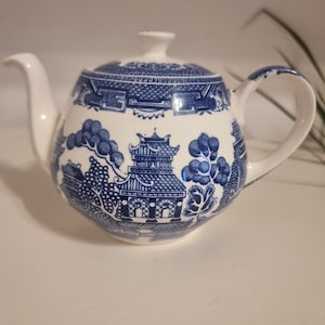 Puede incluir: Una tetera de cerámica blanca con un patrón azul de estilo willow. El diseño incluye ilustraciones detalladas de pagodas, árboles y puentes que rodean el cuerpo. Cuenta con un pico curvo, un asa resistente y una tapa a juego con un pequeño pomo.