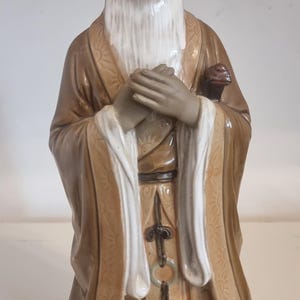 May include: A ceramic figurine of an elderly person with a long white beard and hair, wearing a tan robe with white trim and sunburst patterns. The figure has hands clasped at the chest and a small dark object tucked into the left sleeve area.