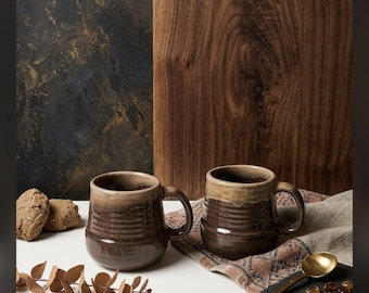 May include: Two ceramic mugs with a dark brown, ribbed lower body and a lighter, smooth tan rim. The mugs feature a rounded handle and a glossy finish. They are arranged with a gold-toned spoon, small brown stones, dried leaves, and cookies.