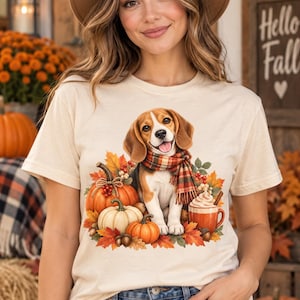 Może przedstawiać: Kremowy t-shirt z grafiką przedstawiającą psa rasy beagle w kraciastej szaliku, otoczonego pomarańczowymi i białymi dyniami, jesiennymi liśćmi, żołędziami oraz kubkiem gorącej czekolady z bitą śmietaną i laską cynamonu.