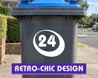 Op de afbeelding: Een donkergrijze afvalbak voor buiten met een felblauw deksel en een opvallende witte cirkelvormige sticker op de voorzijde. De sticker toont het getal 24 in een vetgedrukt, wit lettertype in het midden van een witte ovale vorm.