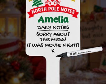Op de afbeelding: Een wit whiteboard met handvat, voorzien van een rode banner met de tekst NORTH POLE NOTES, een groene elf, een boom en gouden sterren. De naam Amelia staat in het groen gedrukt. Inclusief een zwarte whiteboardmarker met tekst WB SL.