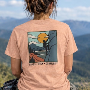 Puede incluir: Una camiseta de manga corta de color melocotón jaspeado con un gráfico cuadrado en la espalda. La ilustración muestra la silueta de una persona escalando una roca frente a un sol amarillo, cielo azul, nubes, montañas y árboles, con el texto ROADS • ROCK • COMMUNITY.