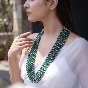 May include: A multi-strand necklace featuring rows of dark green beads interspersed with small white pearls. The necklace is arranged in a layered design, creating a wide, structured collar effect that rests against the chest, secured by a clasp.