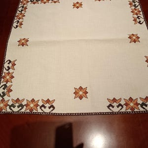 Op de afbeelding: Een vierkante, gebroken witte stoffen doek met een rasterachtige textuur en een kruissteekpatroon. De rand en het midden zijn versierd met herhalende oranje, bruine en crèmekleurige bloemmotieven in geometrische vormen.