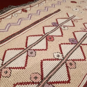 Könnte beinhalten: Ein beigefarbener Stoffläufer mit aufwendiger Kreuzstichstickerei. Das Design umfasst sich wiederholende rote Rauten, kleine rosa und violette Blumenmotive sowie eine lavendelfarbene Zickzack-Borte. Der Stoff weist einen dezenten metallischen Glanz auf.