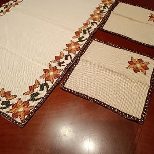 Op de afbeelding: Een rechthoekige tafelloper en twee vierkante placemats van gebroken witte stof met een rastertextuur. De items hebben een kruissteek-randpatroon van oranje en gele bloemen met donkergroene bladeren en een donkerbruine gestikte rand.