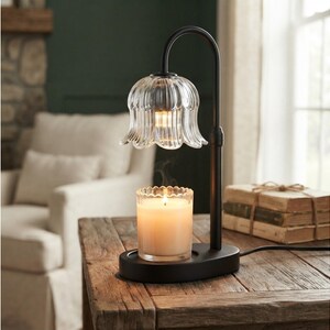 May include: A black metal candle warmer lamp featuring a curved arm and a clear, fluted glass shade shaped like a flower. The lamp sits on an oval base and illuminates a glass jar candle with a visible flame and rising wisps of smoke.