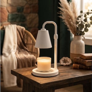 May include: A white candle warmer lamp with a curved neck and a conical shade, featuring a circular base that holds a clear glass jar candle with a glowing wax interior. The lamp has a smooth matte finish and a minimalist structural design.