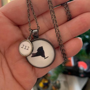 May include: A silver-toned necklace featuring two circular pendants. One pendant displays the silhouette of New York State in black against a white background. The other pendant has the number "212" in black. The necklace is on a silver chain.