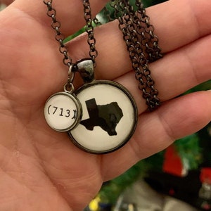 May include: A silver-toned necklace featuring two circular pendants. One pendant displays a black silhouette of Texas on a white background. The other pendant has the area code (713).