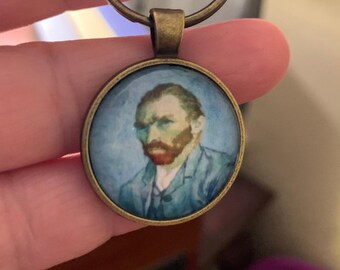 May include: Round keychain with a bronze finish. The pendant features a portrait of Vincent van Gogh in a blue and green color palette. The keychain is held by a hand.