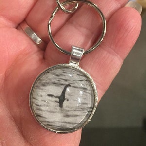 May include: Silver-toned keychain with a round pendant featuring a black and white image of the Loch Ness Monster in a lake. The keychain includes a split ring and a lobster clasp for easy attachment.