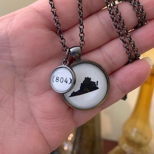May include: A silver-toned necklace with two pendants. One pendant is a circle with the Virginia state outline in black on a white background. The other pendant is a circle with the area code (804) in black on a white background.