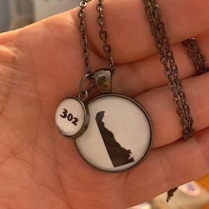 May include: A bronze-toned necklace with two pendants. One pendant features the silhouette of Delaware in black against a white background. The other pendant has the number "302" in black. The necklace is on a chain.