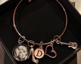May include: A rose gold tone bangle bracelet featuring five hanging charms: a circular pendant with a black and white portrait of a woman, a high-heeled shoe, a round white charm with the letter D, an open heart outline, and an electric guitar.