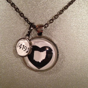 May include: A silver-toned necklace with a pendant featuring a black heart-shaped outline of Ohio. A smaller charm displays the area code (419). The necklace chain is a dark silver color.