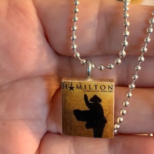 May include: A silver-colored ball chain necklace with a square pendant. The pendant features the word "HAMILTON" with a silhouette of a person in a hat holding a staff. The background is a sepia tone.