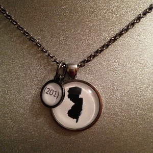 May include: A silver-toned necklace featuring two pendants. One pendant is a circle with the area code (201) in black text. The other pendant is a circle with a black silhouette of the state of New Jersey.