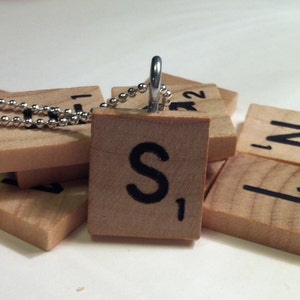 May include: A wooden Scrabble tile pendant with the letter "S" and the number "1" in black. The pendant hangs from a silver ball chain. Other Scrabble tiles are scattered around the pendant.