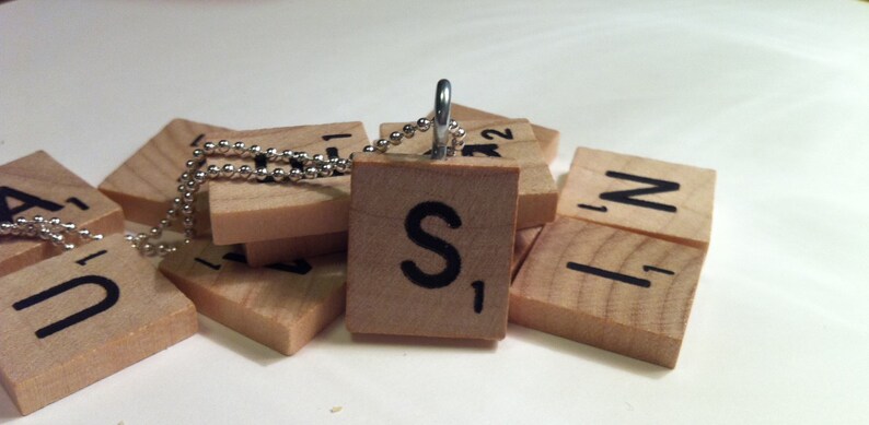 May include: A wooden Scrabble tile pendant with the letter "S" and the number "1" in black. The pendant hangs from a silver ball chain. Other Scrabble tiles are scattered around the pendant.