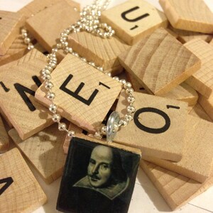 May include: A silver ball chain necklace with a pendant featuring a portrait of William Shakespeare. The pendant is square, set in a wooden frame, and rests on a pile of wooden letter tiles.