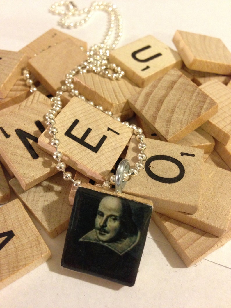 May include: A silver ball chain necklace with a pendant featuring a portrait of William Shakespeare. The pendant is square, set in a wooden frame, and rests on a pile of wooden letter tiles.