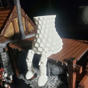 May include: A white 3D printed figure with a geometric honeycomb-patterned torso and articulated legs, sitting on a brown wooden structure. The figure features a hollow top opening and segmented limbs that allow for a seated position on the ledge.