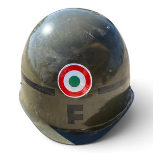 Può includere: Un elmetto militare verde oliva con un emblema circolare a cerchi concentrici rossi, bianchi e verdi centrato sulla parte anteriore. Una striscia orizzontale nera corre lungo i lati e una grande lettera F maiuscola nera è stampata sotto l'emblema.