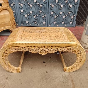 May include: A rectangular wooden coffee table featuring intricate floral carvings across the top surface and along the curved legs. The light-coloured wood displays detailed craftsmanship with scrolling patterns and carved flower motifs throughout the structure.