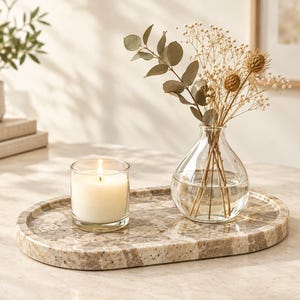 Puede incluir: Una bandeja ovalada de piedra moteada en tonos beige y marrón que sostiene un portavelas de cristal transparente con una vela blanca encendida y un jarrón de cristal transparente con tallos de eucalipto y flores blancas.