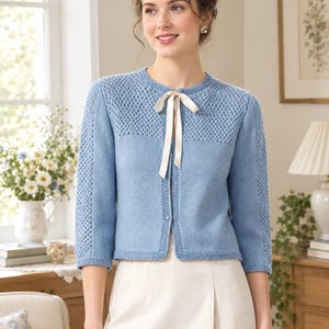 May include: A light blue knit cardigan featuring three-quarter length sleeves with a textured stitch pattern, a matching textured yoke, a round neckline, and a cream-colored ribbon tie closure at the center front. The garment has a ribbed hem.