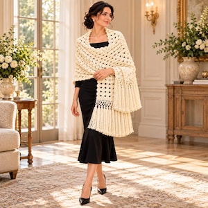 May include: A cream-colored, open-knit crochet shawl featuring a repeating geometric pattern and long, thick fringe along the bottom edge. The shawl is draped over a simple, sleeveless black dress that reaches below the knees.
