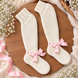 Puede incluir: Un par de calcetines de punto color crema con un puño acanalado en la parte superior y una textura de punto horizontal suave en toda la pierna y el pie. Cada calcetín está adornado con un delicado lazo de satén rosa claro en el tobillo.
