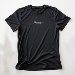 May include: Black crew neck t-shirt with the word "Sacrifice" printed in a gothic font across the chest. The shirt is made of a soft, dark fabric and has short sleeves. The shirt is displayed on a white background.