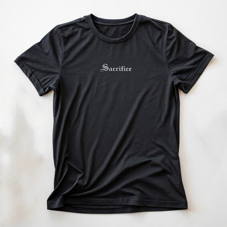 May include: Black crew neck t-shirt with the word "Sacrifice" printed in a gothic font across the chest. The shirt is made of a soft, dark fabric and has short sleeves. The shirt is displayed on a white background.