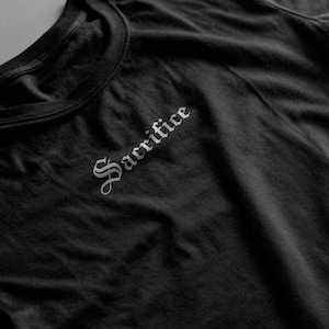 May include: Black crew neck t-shirt featuring the word "Sacrifice" in a gothic font on the upper left chest. The dark fabric of the shirt is visible.