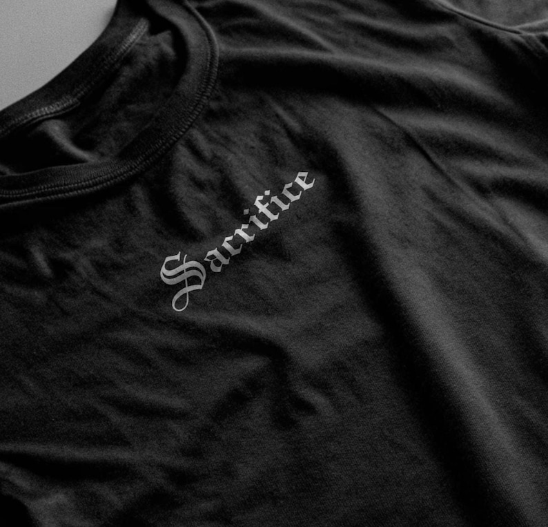 May include: Black crew neck t-shirt featuring the word "Sacrifice" in a gothic font on the upper left chest. The dark fabric of the shirt is visible.