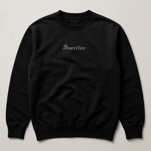 May include: Black crew neck sweatshirt featuring the word "Sacrifice" embroidered in a gothic font. The sweatshirt has a simple, minimalist design, suitable for casual wear.