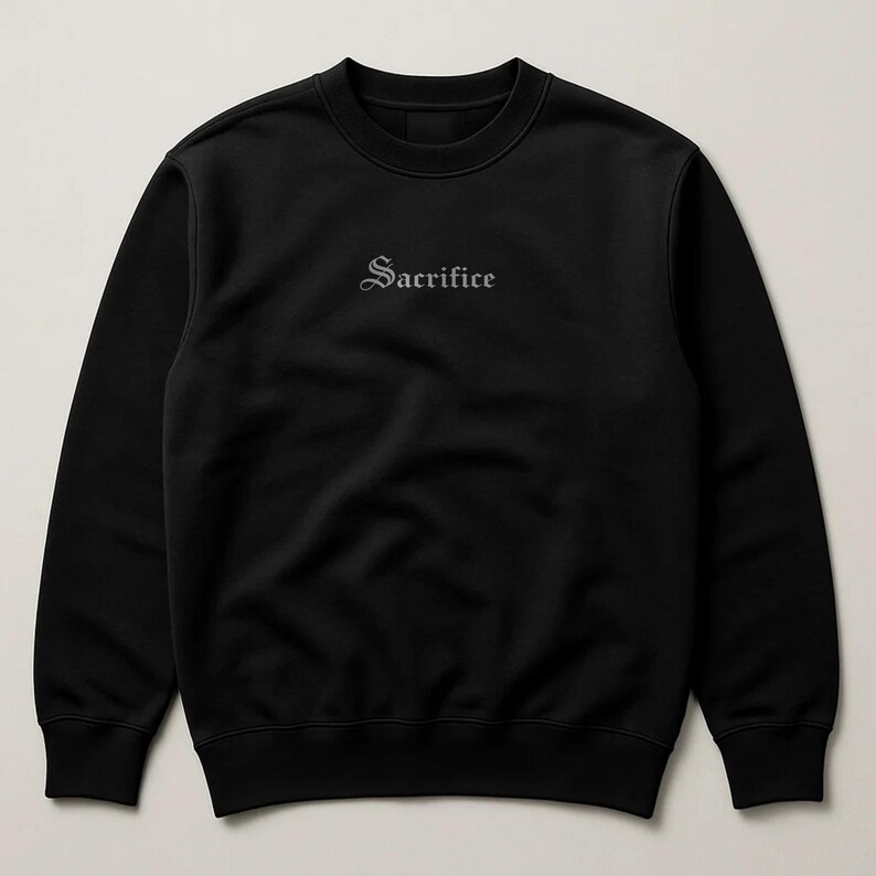 May include: Black crew neck sweatshirt featuring the word "Sacrifice" embroidered in a gothic font. The sweatshirt has a simple, minimalist design, suitable for casual wear.