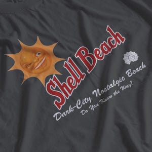 May include: Dark gray t-shirt featuring a winking sun graphic and the words "Shell Beach" in red, with additional text: "Dark-City Nostalgic Beach, Do You Know the Way?" A seashell graphic is also present.