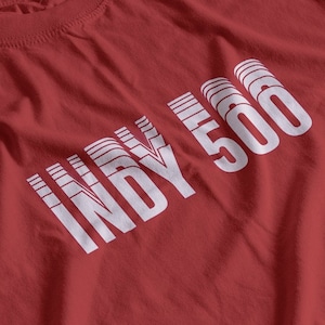 Op de afbeelding: Rood T-shirt met een witte "INDY 500" print op de voorkant. De tekst is in een gelaagde, gestapelde fontstijl.