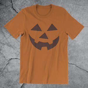 Może przedstawiać: Brązowa koszulka z nadrukiem czarno-białej kratkowanej twarzy jack-o'-lantern.