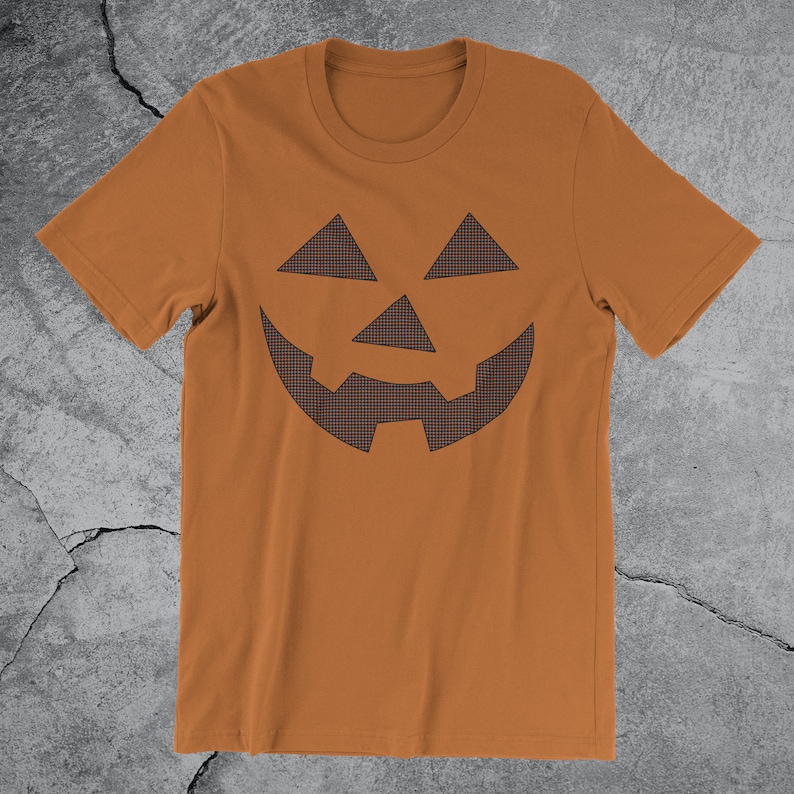 Może przedstawiać: Brązowa koszulka z nadrukiem czarno-białej kratkowanej twarzy jack-o'-lantern.