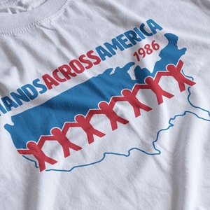 Könnte beinhalten: Weißes T-Shirt mit einer blauen und roten Grafik der Vereinigten Staaten mit dem Text "HANDS ACROSS AMERICA 1986".
